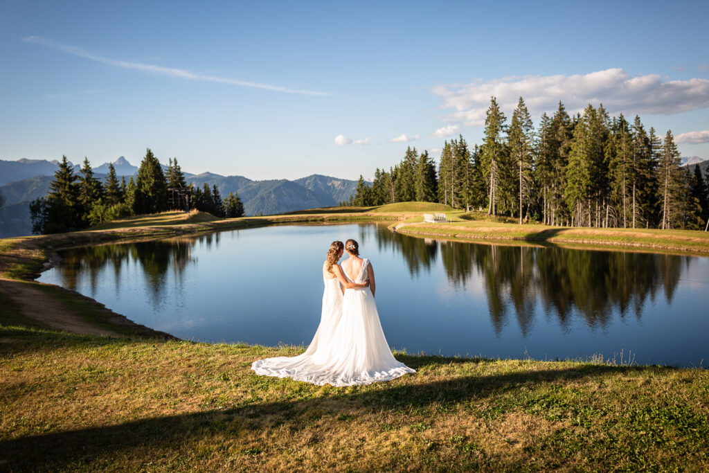 couple de mariées lgbt en robe de mariés devant un paysage magnifique de lac forêt et montagne, photo réalisée par un photographe professionnel spécialisé dans le mariage